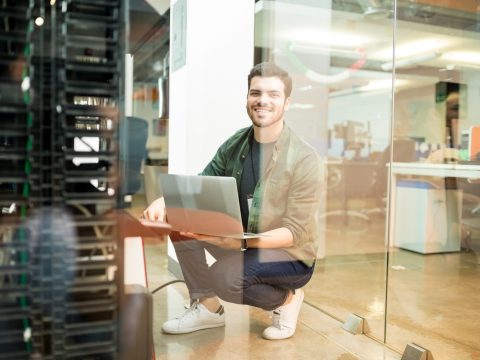 Portrait of happy young male network engineer with laptop in hand working in datacenter.